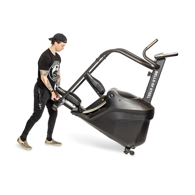 A person dressed in black is adjusting a Bells of Steel Blitz Stepper machine. This cardio equipment, gentle on the joints, offers magnetic resistance for a seamless workout experience against a plain white background.