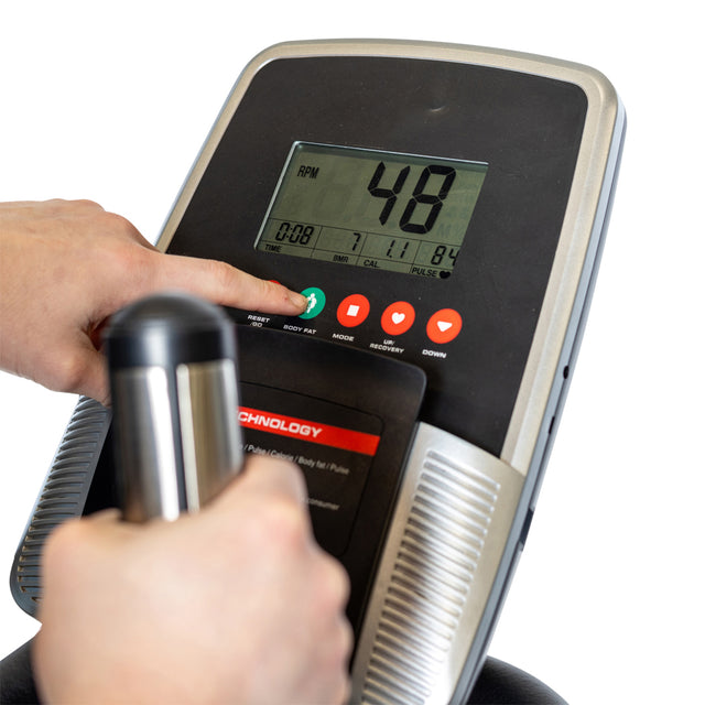 Close-up of a person's hand pressing buttons on the Blitz Stepper's modern display by Bells of Steel, featuring adjustable magnetic resistance. The screen indicates RPM, calories burned, and pulse rate. Another hand grips the handlebar for a joint-friendly cardio workout.