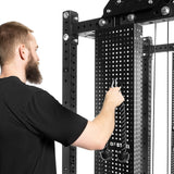 A bearded man in a black shirt adjusts the attachment on a sleek Kraken Weight Stack Shroud from Bells of Steel. The rack-mounted trainer, featuring metal perforated panels and pulleys, ensures home gym safety as he modifies settings.