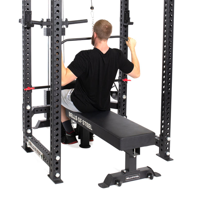 A man in a black shirt sits on a bench doing lat pulldowns with the Lat Pulldown & Low Row Rack Attachment - Hydra & Manticore on a Bells of Steel power rack, pulling down a bar attached to the cable for effective strength training with Olympic Weight Plates.