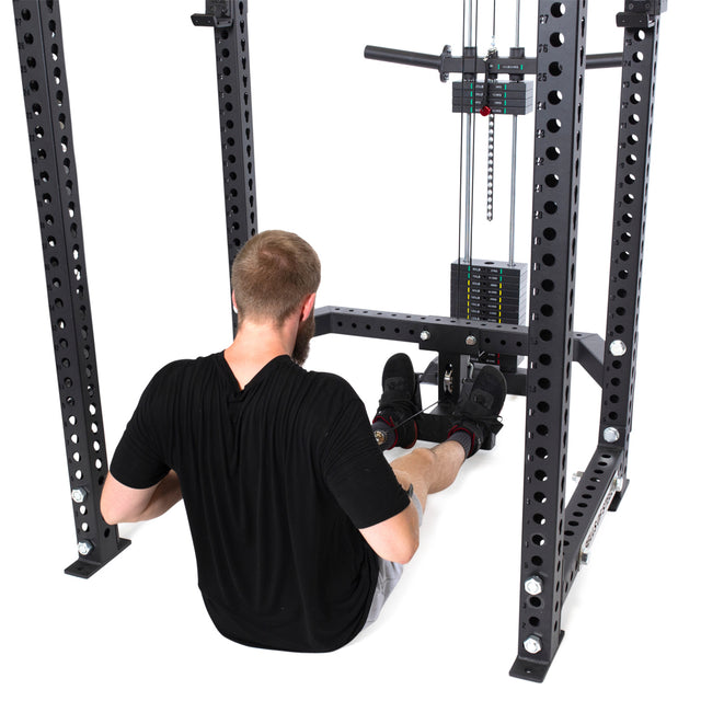 A bearded individual in a black T-shirt and shorts is seated on the gym floor using a Bells of Steel rowing machine with the Lat Pulldown & Low Row Rack Attachment - Hydra & Manticore, enhancing their upper body workout with focused cable exercises.