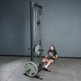 A woman in athletic wear performs seated rows with the Bells of Steel Fat Bar - Straight Handle Bar on a cable machine, aiming to boost grip strength in a gym featuring gray walls and flooring.