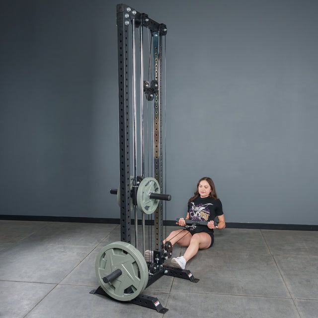 A woman in athletic wear performs seated rows with the Bells of Steel Fat Bar - Straight Handle Bar on a cable machine, aiming to boost grip strength in a gym featuring gray walls and flooring.
