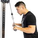 A man in a black t-shirt displays his grip strength using a cable pulley machine equipped with the Bells of Steel Fat Bar - Straight Handle Bar for tricep exercises. He stands sideways, holding the sturdy attachment with both hands against the crisp, white background.