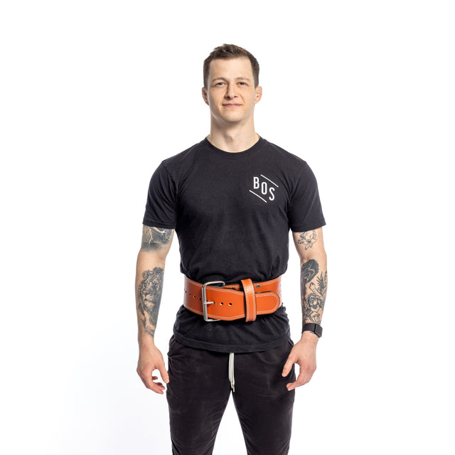 A person confidently wears a black T-shirt featuring "BOS" and displays arm tattoos. They sport a Bells of Steel 10mm Leather Weightlifting Belt with a large brown single-prong in front of a plain white background, accentuating their bold style.