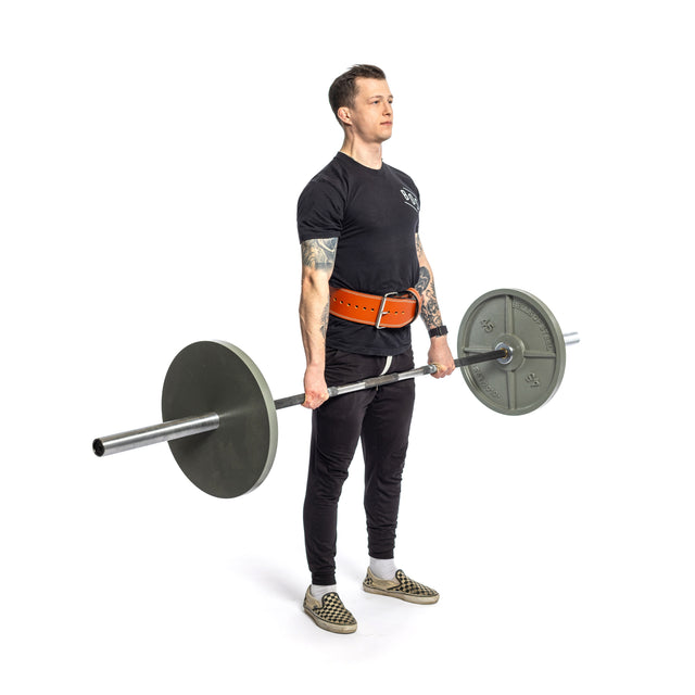 A person in a black shirt and pants performs a deadlift with a barbell, sporting a Bells of Steel 10mm Leather Weightlifting Belt and patterned slip-on shoes, set against a plain white background.