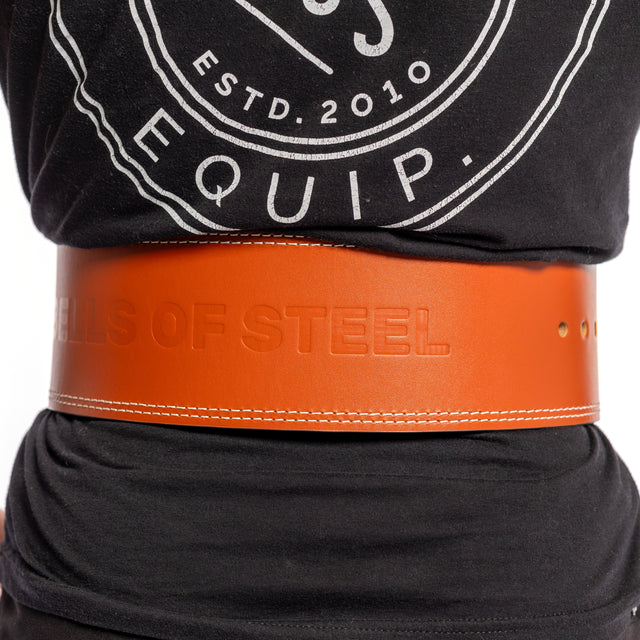 A person in a black shirt with a circular logo wears a wide brown 10mm Leather Weightlifting Belt by Bells of Steel, featuring an embossed "BELLS OF STEEL" text. It is securely fastened around the waist with its single-prong buckle.