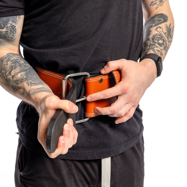 A person tightens a sturdy Bells of Steel 10mm Leather Weightlifting Belt around their waist. Sporting a black t-shirt, they showcase their arm tattoos, while the belt's vibrant orange color and silver buckle perfectly complement their weightlifting look.