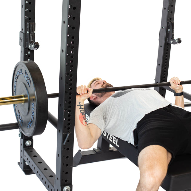 Using the Bells of Steel Sandwich J-Cups for support, a person performs a bench press on a gym bench with weights on a barbell. They are wearing a light gray shirt and black shorts, gripping the bar firmly while HDPE padding ensures maximum comfort and stability.