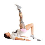 A tattooed individual uses Bells of Steel Core Sliders to perform a single-leg glute bridge, lying on their back with one leg raised and arms flat, dressed in a gray T-shirt and pink shorts for a total body workout.