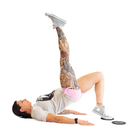 A tattooed individual uses Bells of Steel Core Sliders to perform a single-leg glute bridge, lying on their back with one leg raised and arms flat, dressed in a gray T-shirt and pink shorts for a total body workout.