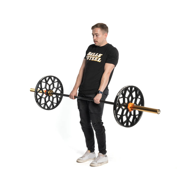 A man in a black "Bells of Steel" T-shirt and dark jeans lifts a barbell with Bells of Steel Technique Plates - 5 Lbs (Pair). He stands on a white background, wearing white sneakers, appearing focused and embodying the intensity of a home gym session.