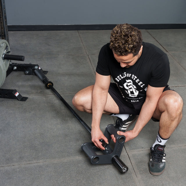 A person in a black Bells of Steel t-shirt and shorts kneels on the gym’s gray floor, attaching a barbell to a landmine. The Bells of Steel Fat Bar - T-Bar Row Cable Attachment rests nearby against the gray wall.