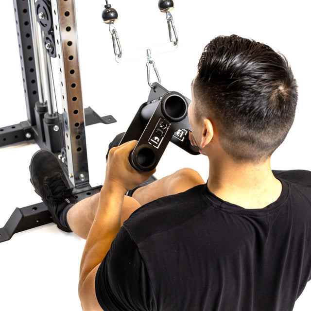 A person with short dark hair uses the Bells of Steel Fat Bar - T-Bar Row Cable Attachment for seated rows at the gym, improving grip strength. They're in a black shirt, holding the bar attached to a cable machine, with visible frame and pulleys behind them.