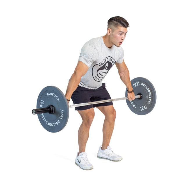 A person in a gray T-shirt and black shorts focuses intensely on their lifting skills, performing a deadlift with the Bells of Steel Aluminum Technique Barbell in a gym. They're also sporting white sneakers.