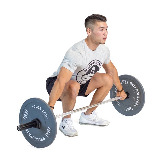 A person is preparing to lift a loaded Aluminum Technique Barbell from Bells of Steel in the gym, sharpening their skills. They are wearing a gray T-shirt, dark shorts, and white sneakers.