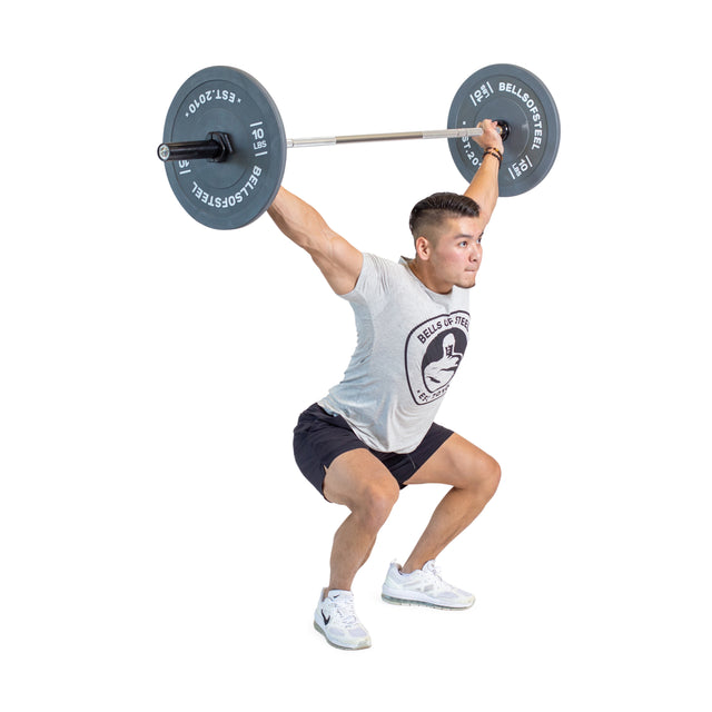 Wearing a gray T-shirt and black shorts, a person demonstrates impressive lifting skills with an Aluminum Technique Barbell by Bells of Steel, performing an overhead squat. Positioned sideways against a white background, they exhibit focus and strength during the exercise.