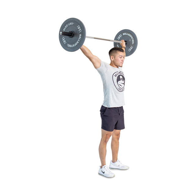 A person expertly lifts a Bells of Steel Aluminum Technique Barbell overhead, wearing a gray T-shirt, black shorts, and white sneakers against a plain white background.