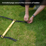 A person anchors the Agility Ladder and Leg Cord Bundle from Bells of Steel to grassy ground using black straps, a yellow level tool, and included pegs, demonstrating how each ladder corner can be secured for stability during agility training. Explanatory text is shown above describing this feature.