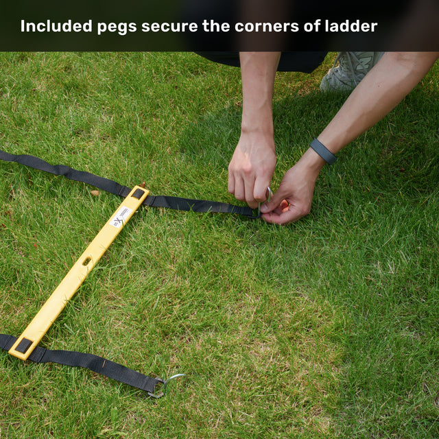 A person anchors the Agility Ladder and Leg Cord Bundle from Bells of Steel to grassy ground using black straps, a yellow level tool, and included pegs, demonstrating how each ladder corner can be secured for stability during agility training. Explanatory text is shown above describing this feature.
