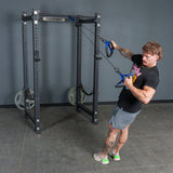 A person performs a standing row exercise using the Bells of Steel Rotational Pulley attached to a power rack in a gym, pulling the handles toward their body.