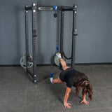 A person performs a suspended plank in a gym setting with their feet secured in harnesses attached to cables on a squat rack, utilizing the Bells of Steel Rotational Pulley for added resistance, while keeping their hands on the floor.