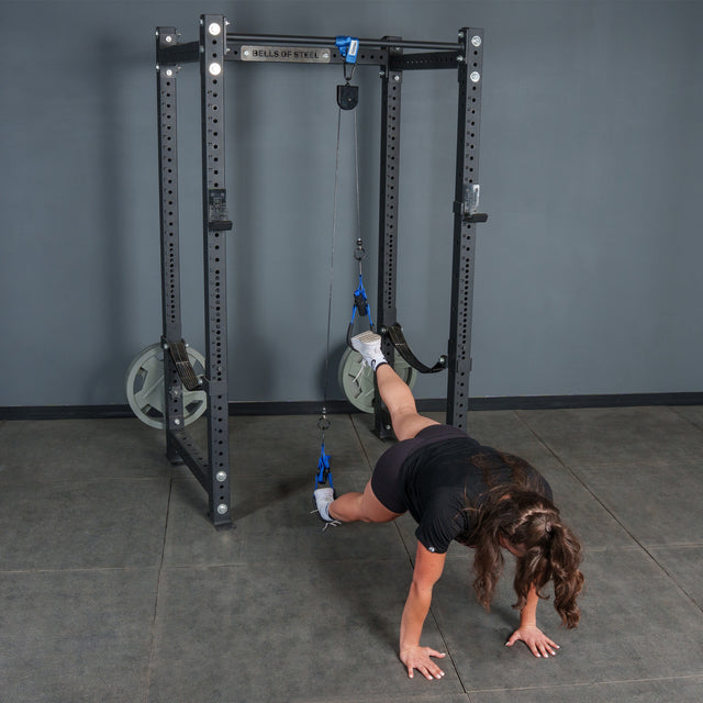 A person performs a suspended plank in a gym setting with their feet secured in harnesses attached to cables on a squat rack, utilizing the Bells of Steel Rotational Pulley for added resistance, while keeping their hands on the floor.