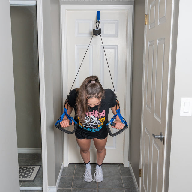 A person uses the Bells of Steel Rotational Pulley, attaching it to a door frame and gripping the handles for a bodyweight workout, exercising in a hallway while wearing a graphic T-shirt, shorts, and sneakers.