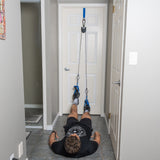 A person lies face up on the floor in a home hallway, performing a bodyweight workout with both feet secured in the Bells of Steel Rotational Pulley system, which is attached to the top of a closed door.
