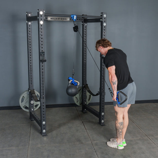 Wearing athletic attire, an individual performs a triceps exercise in a gym with grey walls and rubber flooring, using the Bells of Steel Rotational Pulley with a kettlebell attached.