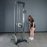 Wearing athletic wear in a gym with gray walls and concrete floors, a woman uses the Bells of Steel Fat Bar - Tricep Pressdown, gripping the handles with both hands to perform a triceps pushdown and build grip strength.