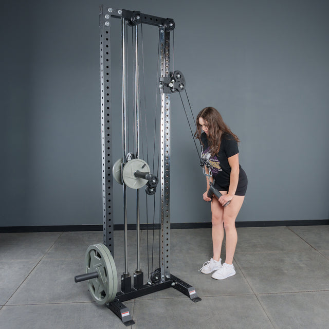 Wearing athletic wear in a gym with gray walls and concrete floors, a woman uses the Bells of Steel Fat Bar - Tricep Pressdown, gripping the handles with both hands to perform a triceps pushdown and build grip strength.