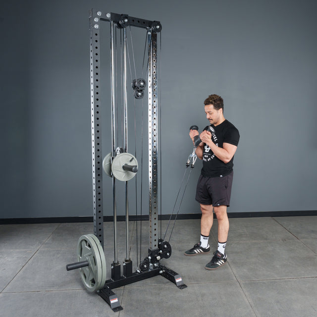 A man in a black t-shirt uses the Bells of Steel Fat Bar - Tricep Pressdown on a cable pulley in a gym with gray walls and concrete floors, focusing on grip strength during the exercise.