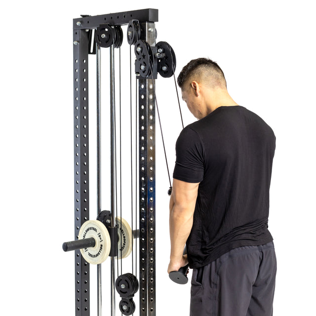 At a bustling gym, a man wearing a black shirt and gray pants works out on the Fat Bar - Tricep Pressdown from Bells of Steel. Facing the machine, he focuses on strengthening his triceps with each pull of the handlebar attached to the weights, his back turned toward those observing him.