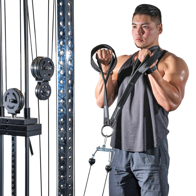 A muscular person is using the RocIT Tricep Strap from Bells of Steel at the gym to perform an arm exercise. They wear a sleeveless black top and dark shorts, focusing intently on targeting their triceps. The machine features adjustable weights and pulleys for optimal resistance.