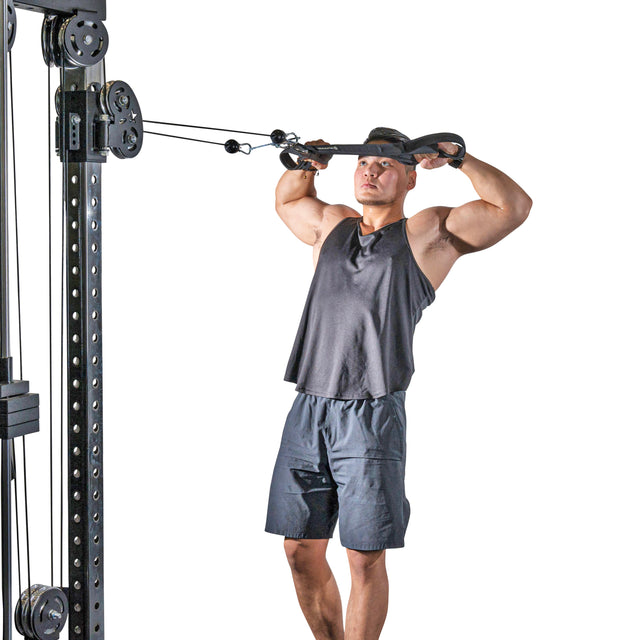 A man wearing a black tank top and shorts is at the gym, using a RocIT Tricep Strap from Bells of Steel to perform a cable face pull exercise. He grips the attachment with both hands, pulling it toward his face with intense focus, while the white background accentuates his tricep targeting.