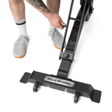 A person in gray sneakers and white socks adjusts a handle on a black Bells of Steel Adjustable Utility Bench with front wheels and 575lb capacity, set against a white background.