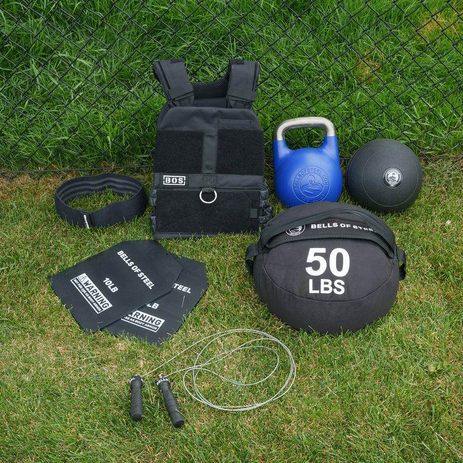 The Ultimate Backyard Training Bundle by Bells of Steel—featuring a weight vest, kettlebell, medicine ball, sandbag, jump rope, lifting belt, and steel plates—arranged on grass in front of a chain-link fence. This complete set is ideal for outdoor workouts and building your own backyard gym with essential fitness equipment.