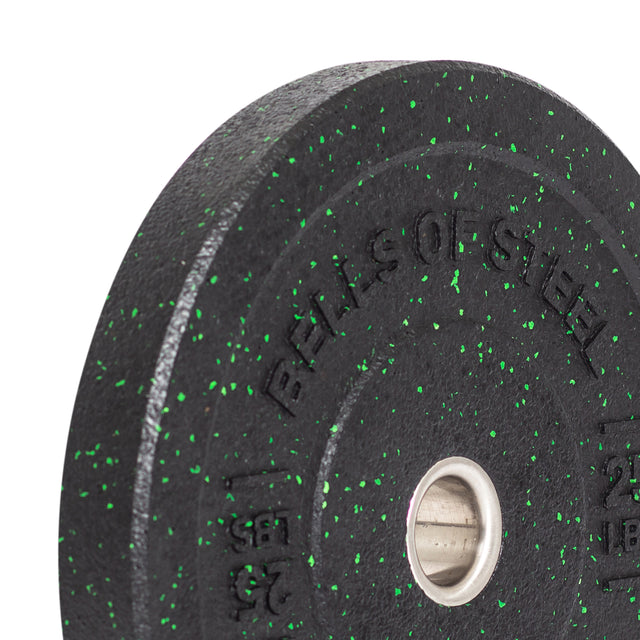 A close-up shot of a Bells of Steel Crumb Bumper Plate, black with green speckles, showcases "BELLS OF STEEL" engraved on it and includes a stainless steel insert ring for barbell attachment.