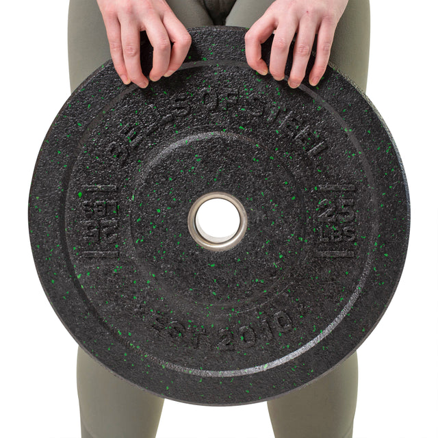 A person is holding a 25 lbs black crumb bumper plate with green specks, featuring a stainless steel insert ring, made by Bells of Steel. It complements their olive green leggings while their face stays out of view.