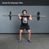A person stands in a gym holding the Bells of Steel Multi-Purpose Olympic Barbell – The Utility Bar at shoulder height, ready to lift. Text above reads, "Great for Olympic lifts." They wear a graphic tee, shorts, and athletic shoes.