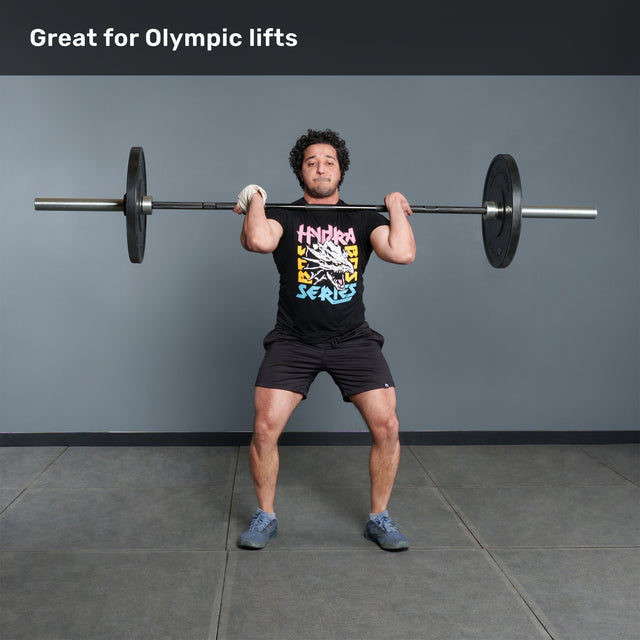A person stands in a gym holding the Bells of Steel Multi-Purpose Olympic Barbell – The Utility Bar at shoulder height, ready to lift. Text above reads, "Great for Olympic lifts." They wear a graphic tee, shorts, and athletic shoes.