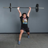 Wearing a black graphic tee, shorts, and athletic shoes, a person with one arm bandaged stands in a gym split stance, performing an overhead lift using the Bells of Steel Multi-Purpose Olympic Barbell – The Utility Bar.