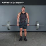 A muscular man in a gym holds the Bells of Steel Multi-Purpose Olympic Barbell – The Utility Bar, wearing a black tank top, shorts, and a weightlifting belt. Text above reads “1000lbs weight capacity.”.