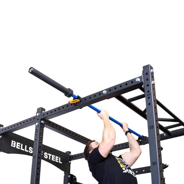 A person is performing a pull-up on a blue Above Rack Barbell Holder, securely set on the black Bells of Steel pull-up station. The sturdy power rack features metal beams and bolts, all against a plain white background.