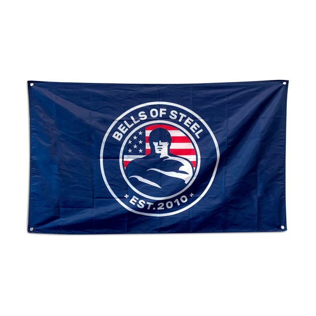 A blue flag from Bells of Steel features a circular emblem depicting a muscular figure standing in front of a stylized American flag. Surrounding the image are the words "BELLS OF STEEL EST. 2010," bringing an inspiring accent to your gym decor or home workout space.