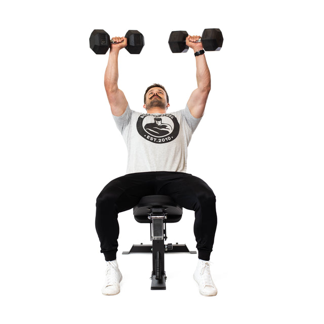 Seated on the Bells of Steel Adjustable Utility Bench, a person lifts two dumbbells in an overhead press. They wear a gray t-shirt, black pants, and white sneakers against a plain white background.