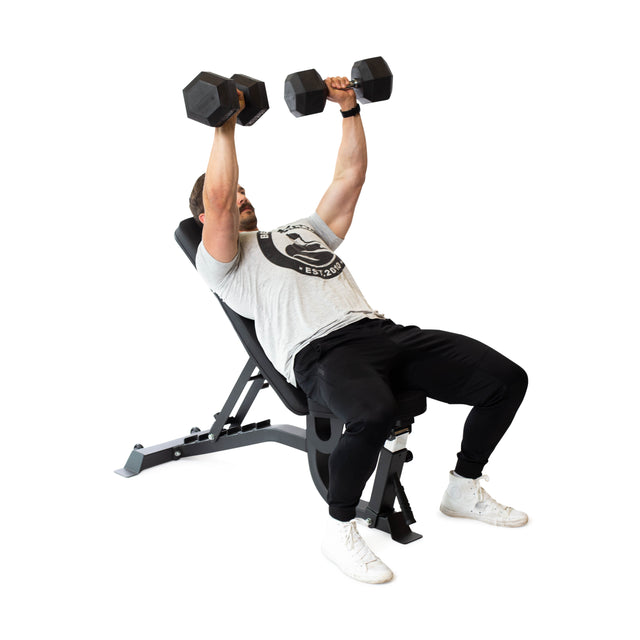 A person performs a dumbbell incline bench press on the Bells of Steel Adjustable Utility Bench, dressed in a gray t-shirt, black pants, and white sneakers, lifting weights against a plain white background.