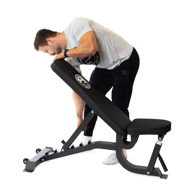 A person in a gray shirt and black pants adjusts the incline angles of the Bells of Steel Adjustable Utility Bench with its metallic frame on a white background.