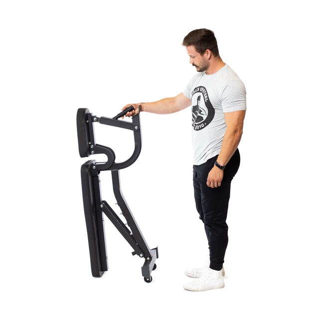 A person in a gray t-shirt and black pants demonstrates the portability of the Bells of Steel Adjustable Utility Bench, with its black color, wheels, and customizable incline angles, against a white background.